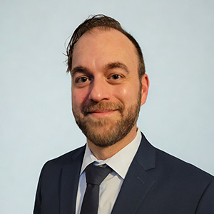 A man in a black suit and tie poses in front of a light blue background