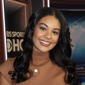 A woman in a brown sweater is seen in front of a TV studio set