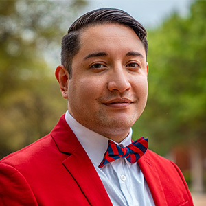 A man in a red suit and bow tie is seen in front of a row of trees