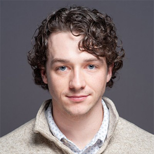 A man in a beige sweater is seen in front of a gray background