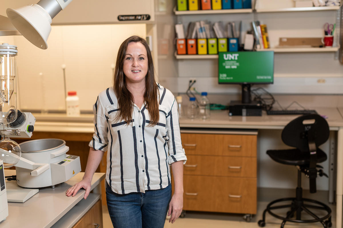 A woman in a white blouse with black stripes and jeans stands in a lab with a smile.