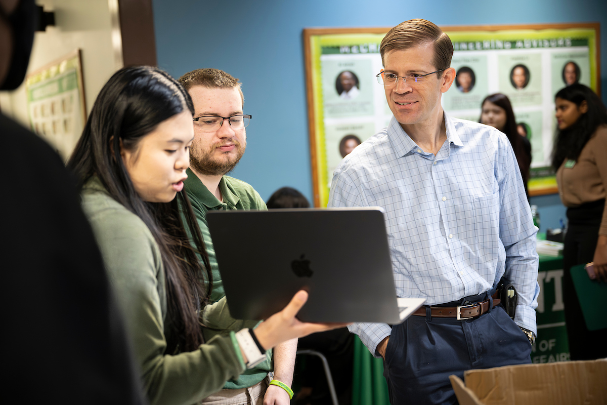 A man in a button down shirt on the right looks at a laptop held up by a student on the left.