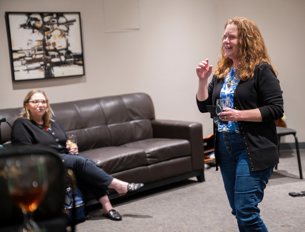 Watkins stands in a room speaking. She holds a card and glass. A woman sits on a sofa out of focus listening.
