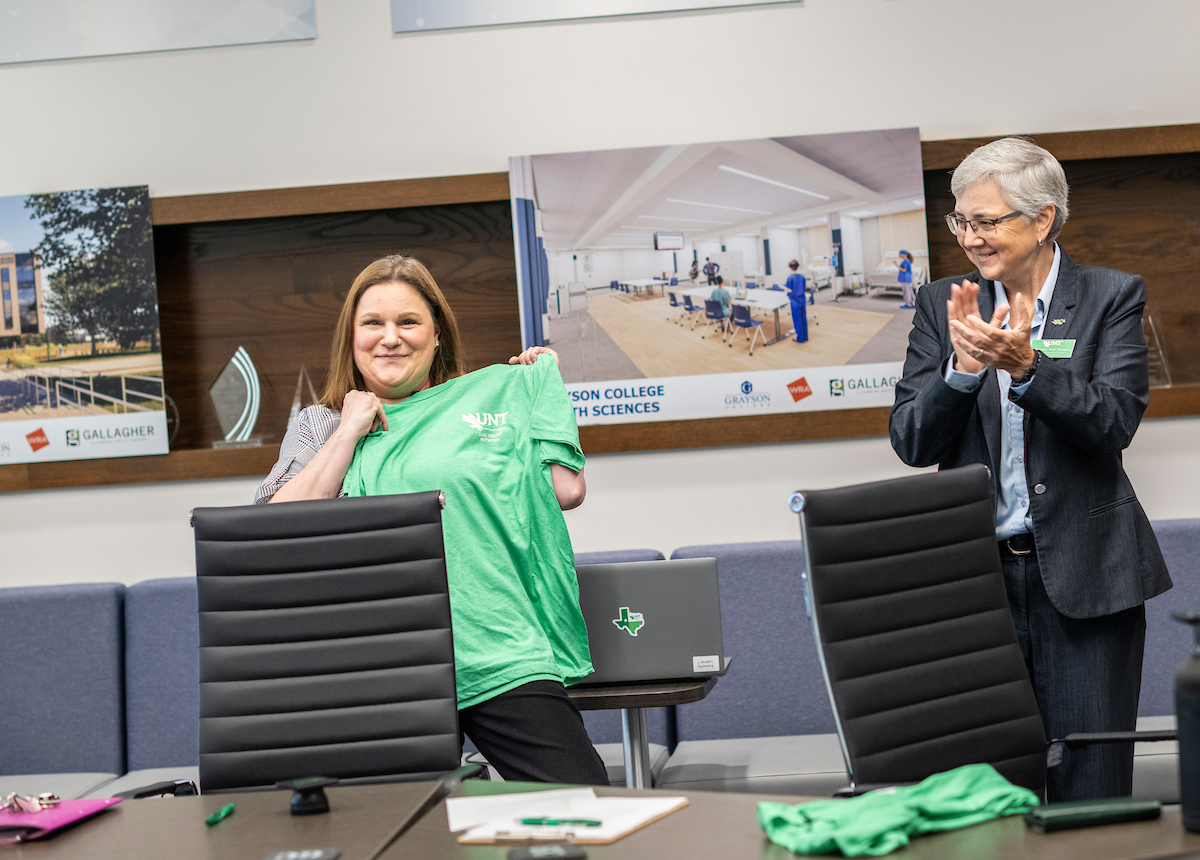 A photo of two women. One stands holding up a green UNT shirt while smiling. The second applauds her.