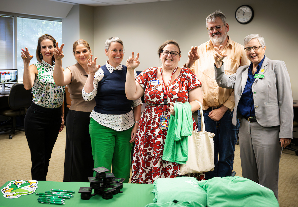 A group of five women and a man stand in a line making the eagle claw sign at the camera. In front of them is a green table with UNT swag on it.