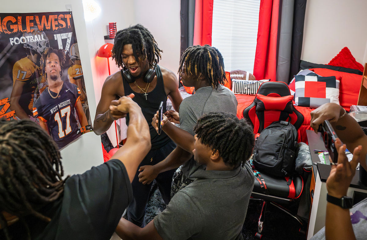 A young man in a black tank top celebrates his new room with three os his friends surrounding him. His rooms red, black, and grey and features a poster of a basketball player.