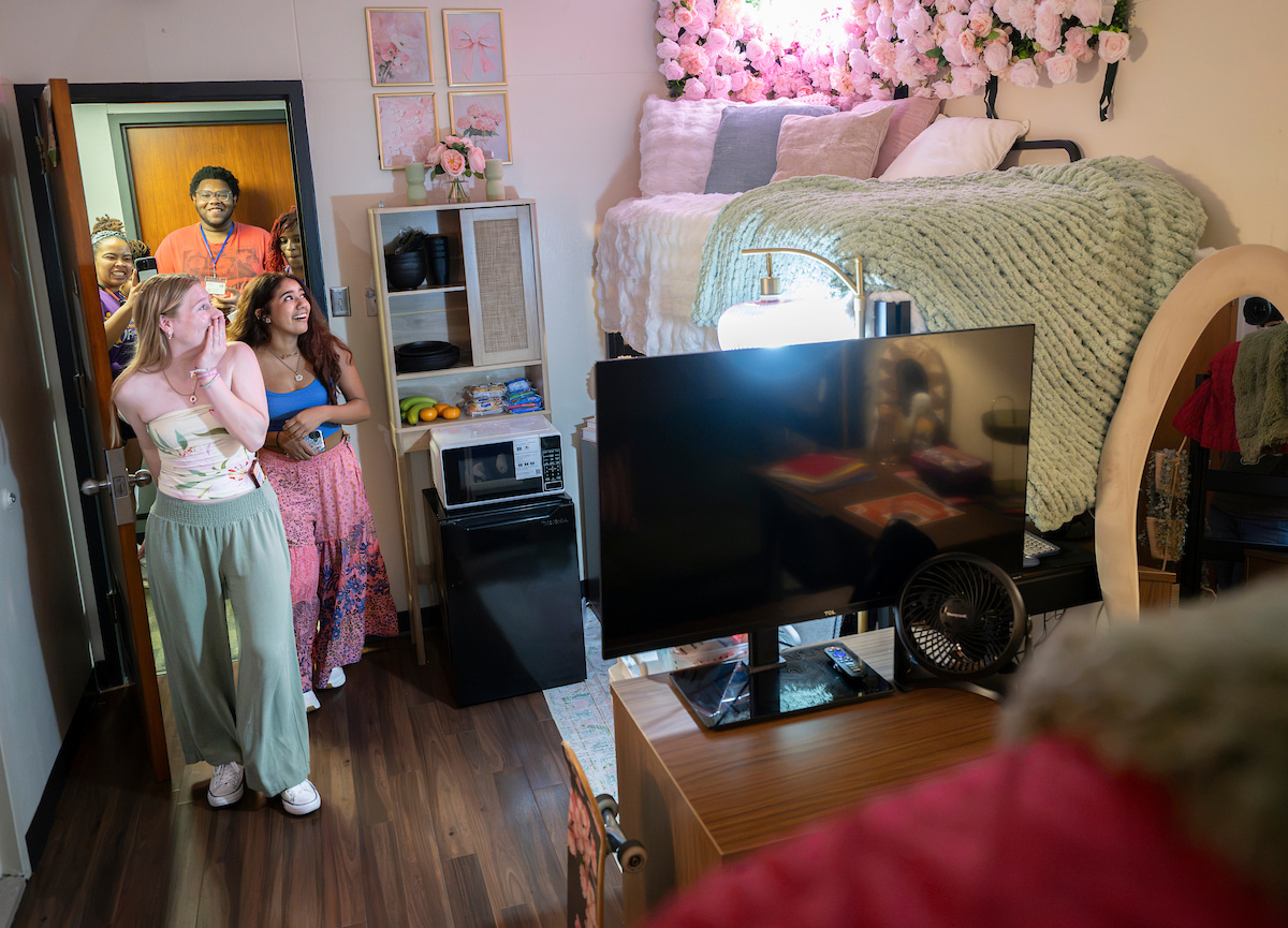 Two young women walk into their dorm with a shocked expression. They look to their right at a lifted dorm bed surrounding by pink flower wall art.