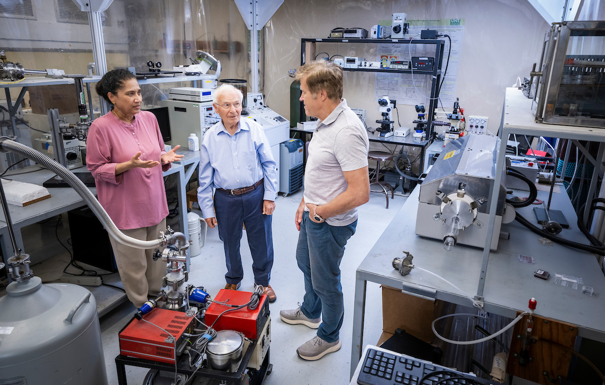 A woman in a pink blouse speaks to two men wearing a light blue button down and gray polo respectively. They all stand in a physics lab surrounded by equipment.