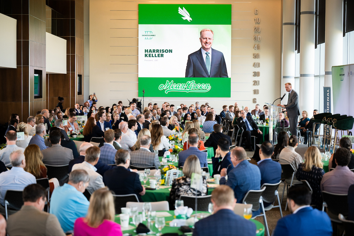 A crowd sits at tables looking up at a man at a podium. In the back is a projector that reads Harrison Keller with his photo.