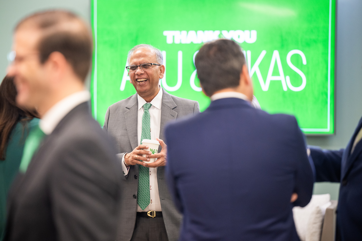 The camera focuses on a smiling man in a suit. Other people walk around him.