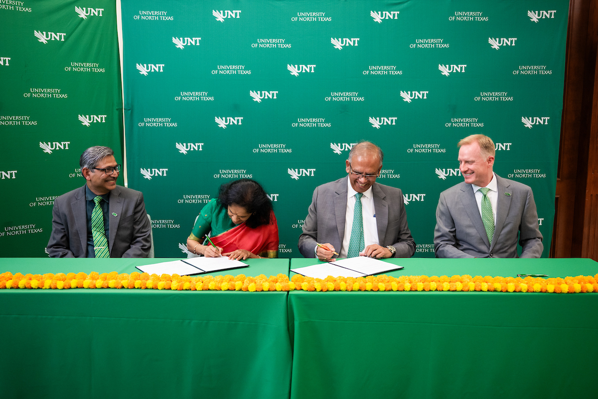Four people, three men one woman, sit at a green table smiling at the camera