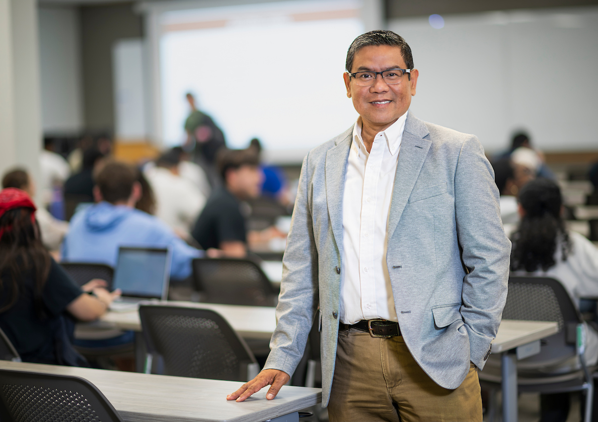 A man in a light gray blazer, button down, and khaki pants smiles at the camera. He is in a full classroom.