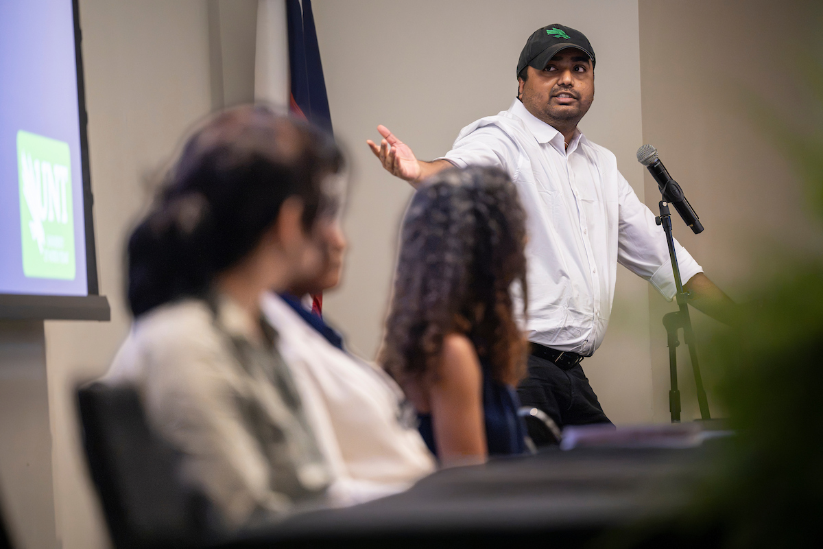 A man in a white button down and black hat speaks into a mic and gestures towards a screen in the corner of the photo.