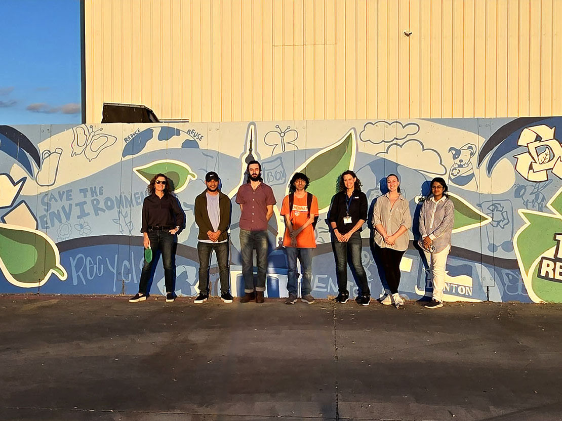 Professor Jamie Johnson and students pose in front of a colorful mural at the Pratt Recycling Facility in Denton, Texas