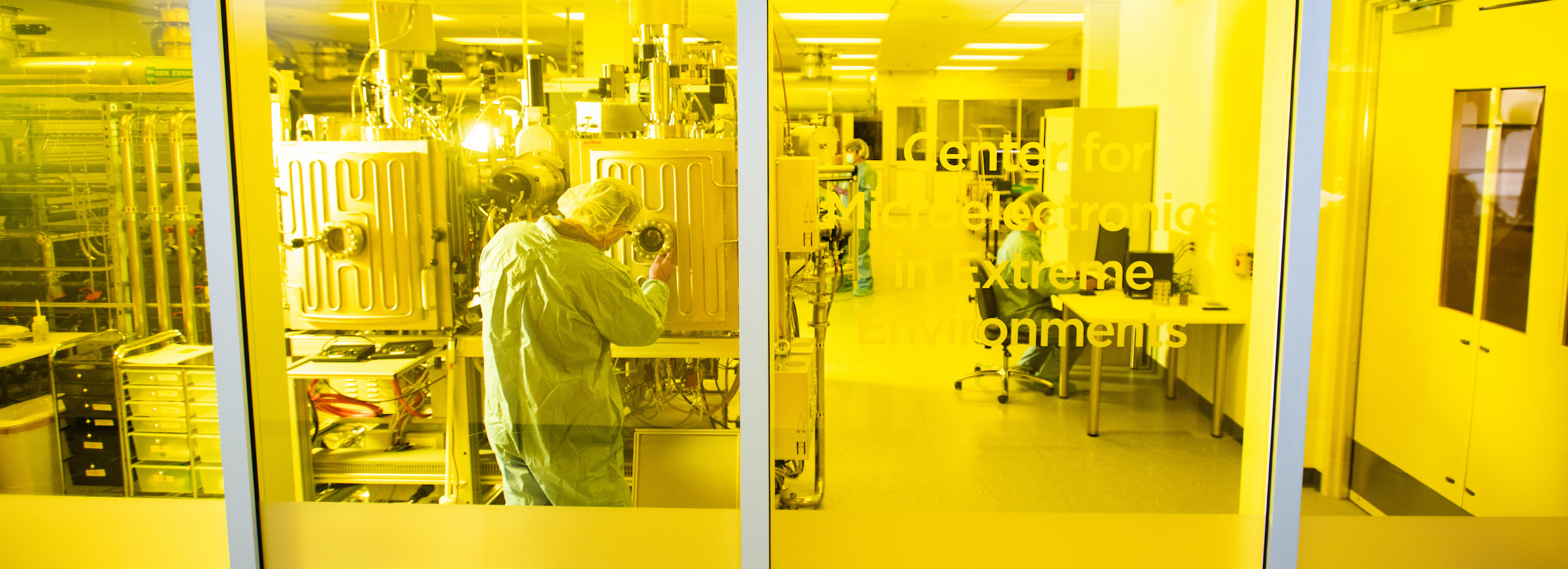 A photo of a lab behind yellow tinted windows. A person in full scrubs and hairnet attends a large machine while another works on a computer