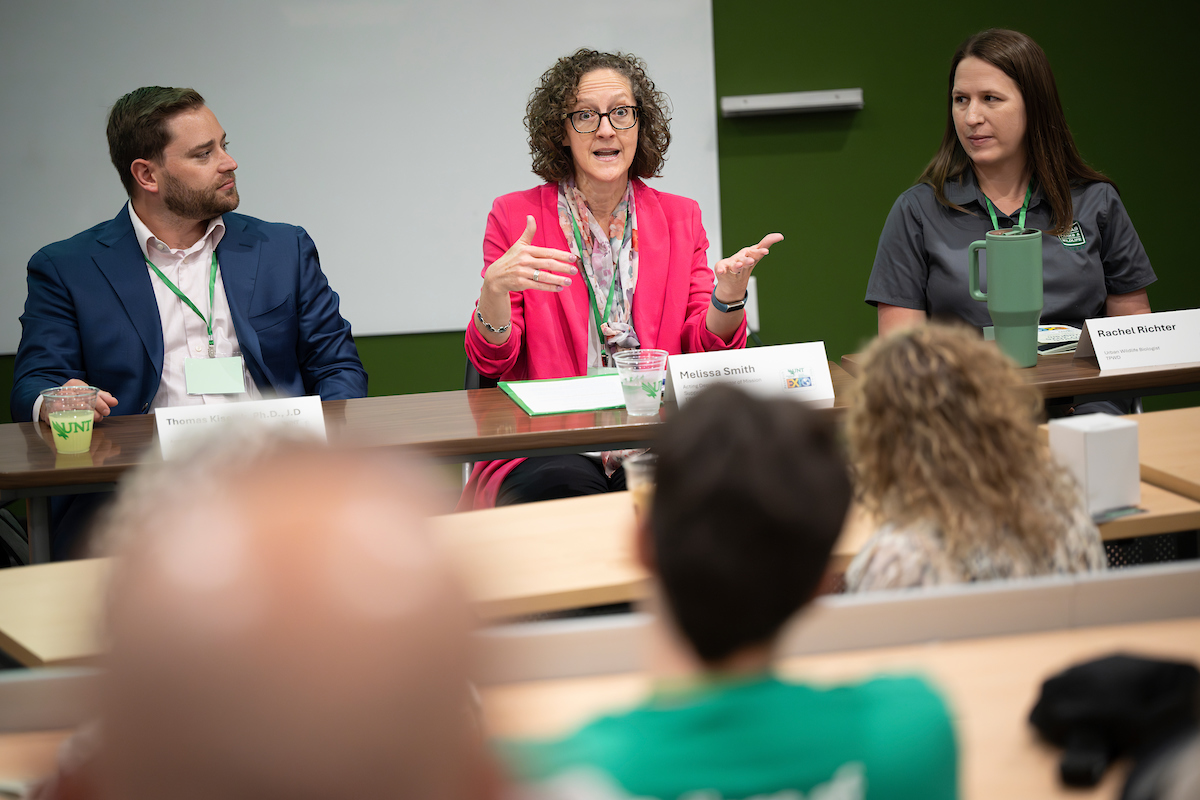 a group of onlookers look at the three panelists, one man and two women, the woman in the center is talking