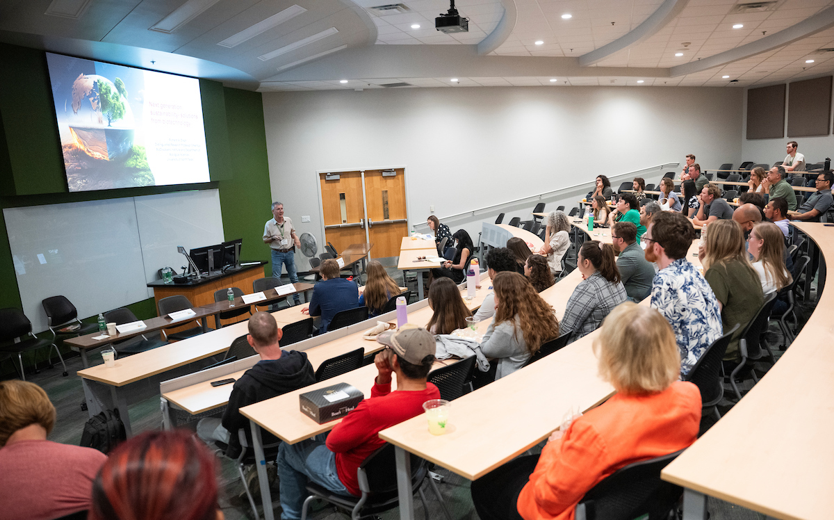 A large group of people sit at desks and watch a presentation from Richard Dixon