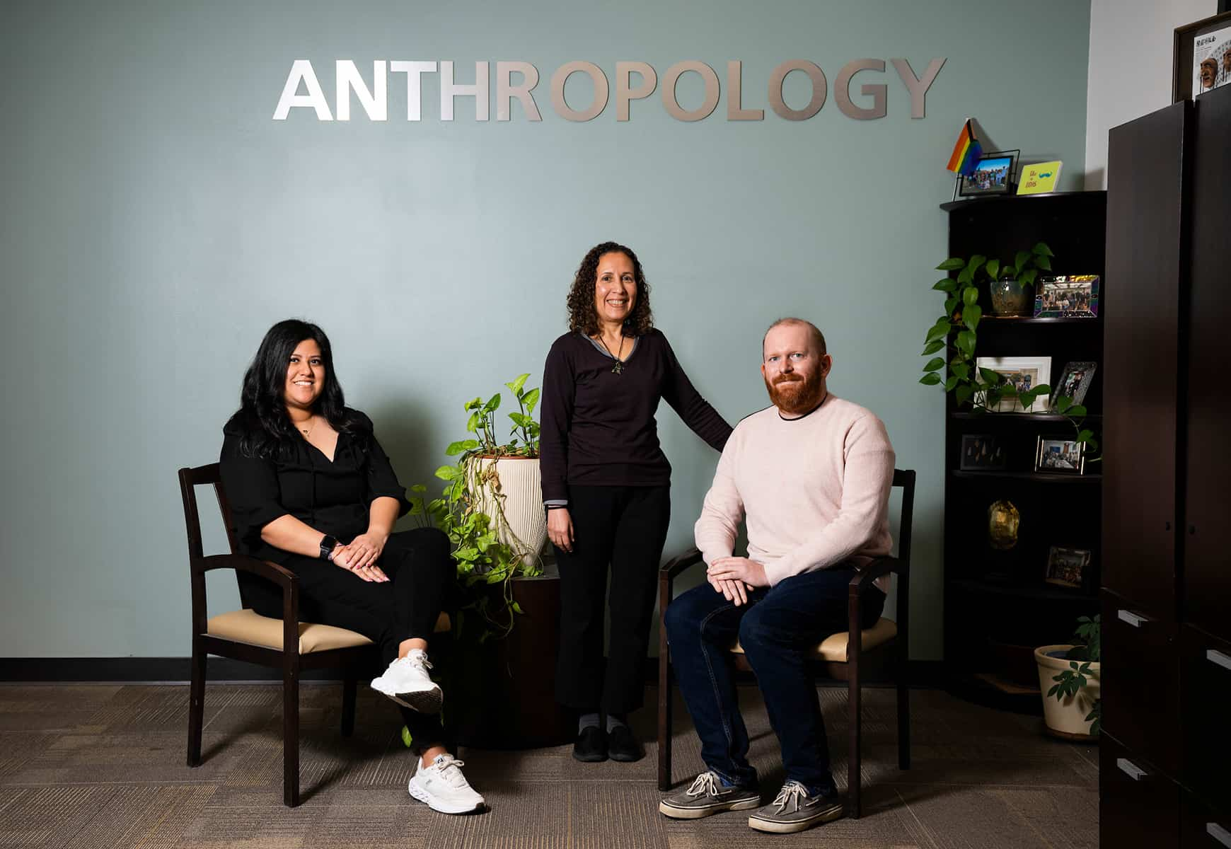 Two students sit while a professor stands behind them; behind all three is a wall with a plaque reading "Anthropology"