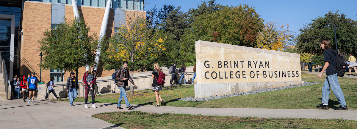 Students walk outside of a large brick building with a sign that says G. Brint Ryan College of Business