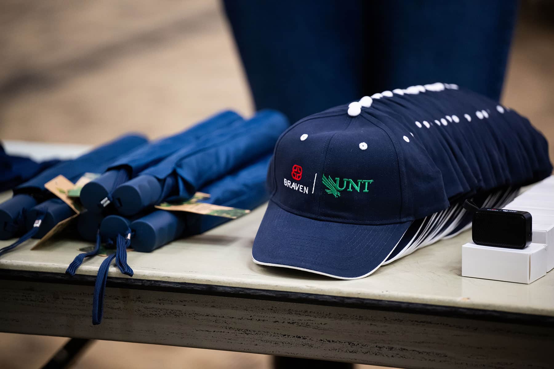 Navy blue caps with the Braven and UNT logos sit in a line on a table next to navy umbrellas