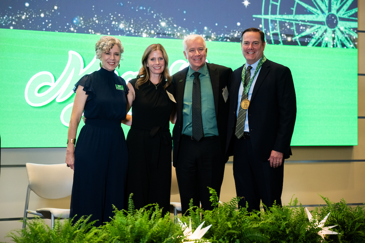 Two women and 2 men stand side by side posing for a photo. The women wear all black while the men wear green shirts with black pants and blazer. The man on the far right, Brian Richardson, wears an award medal