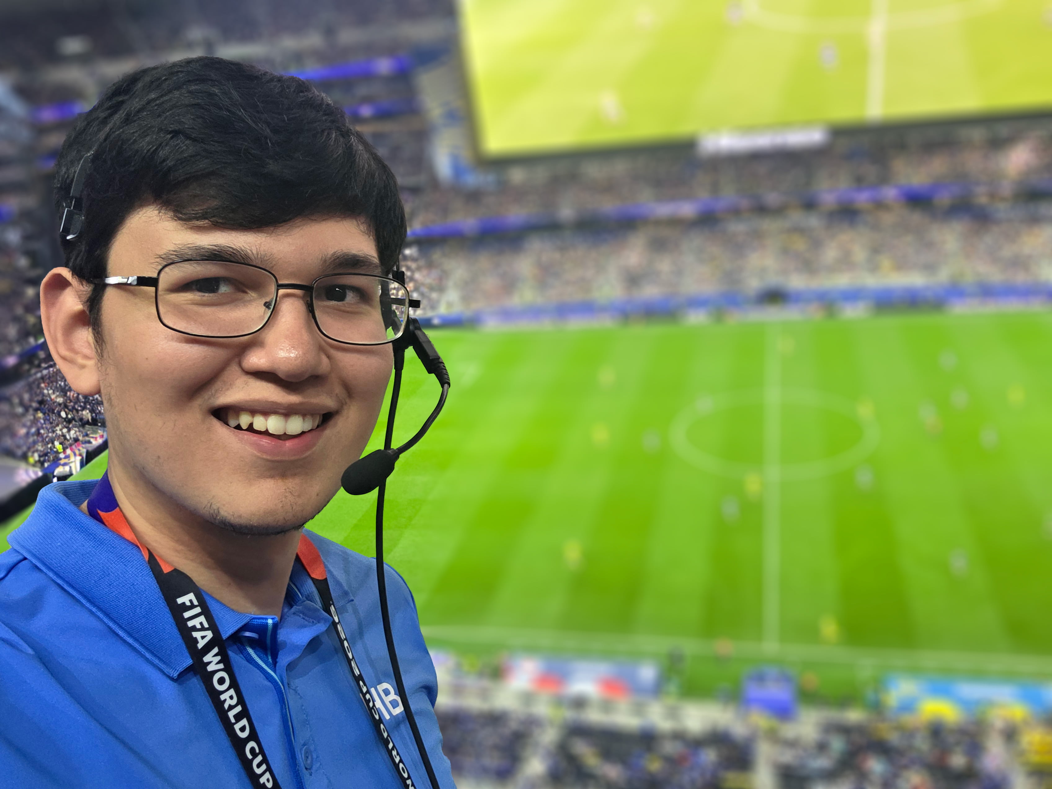 A selfie of a young man in a blue polo and production headset in a stadium with a soccer field below him