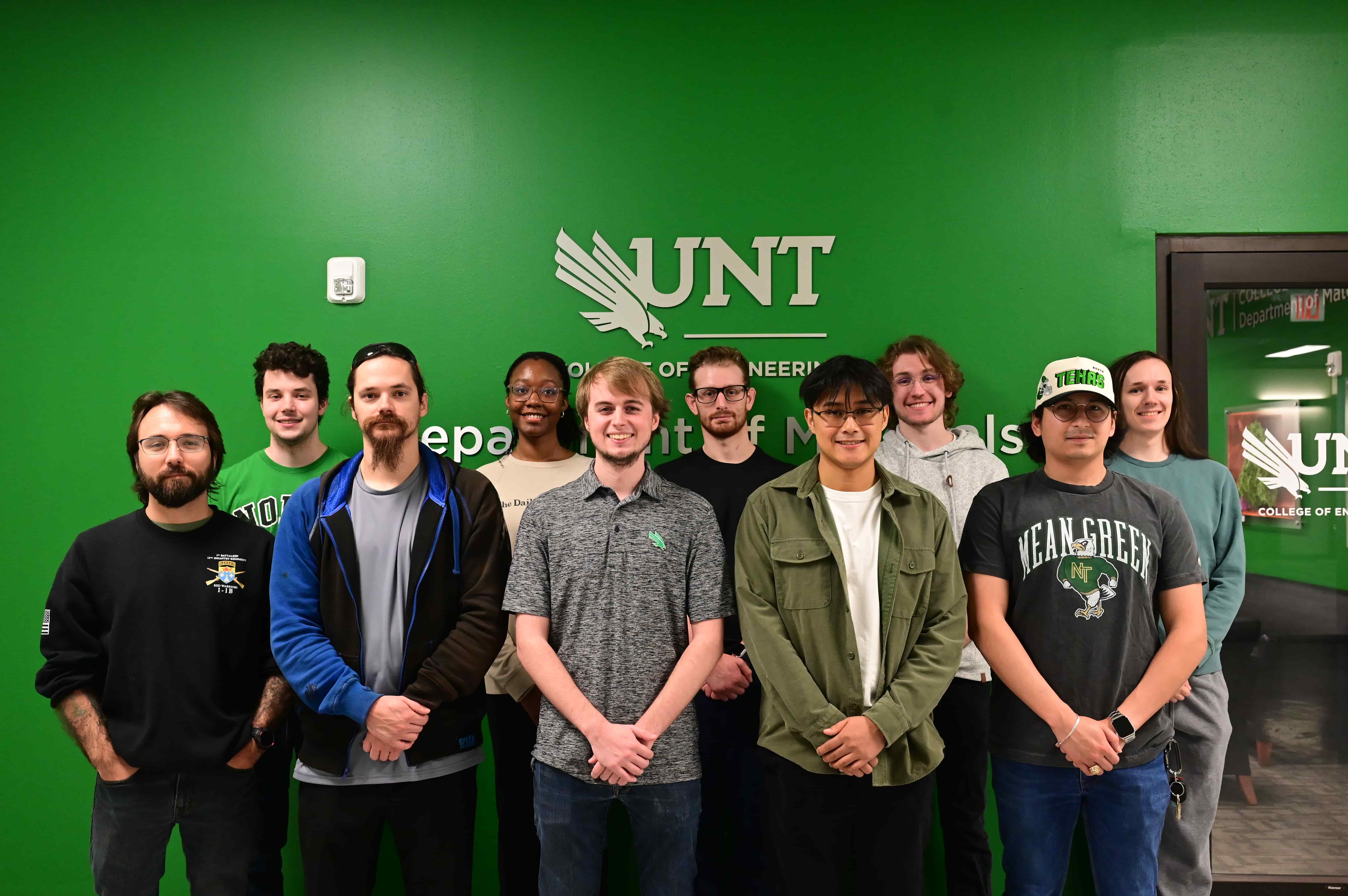A group of 12 students of stand in front of a green wall with the letter UNT on it. Most clasp their hands in front of them.