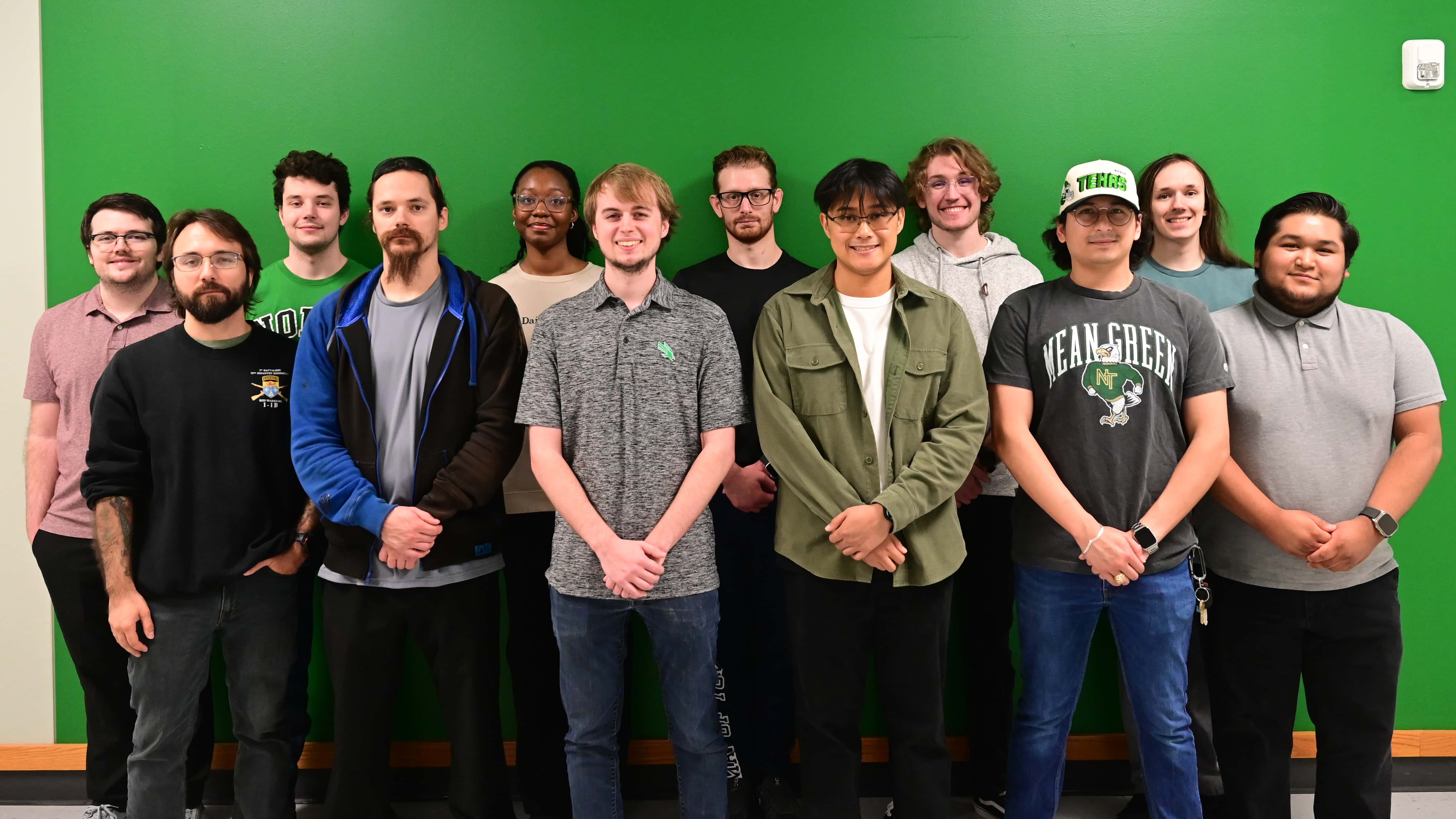 A group of 12 students of stand in front of a green wall. Most clasp their hands in front of them and smile are the camera.