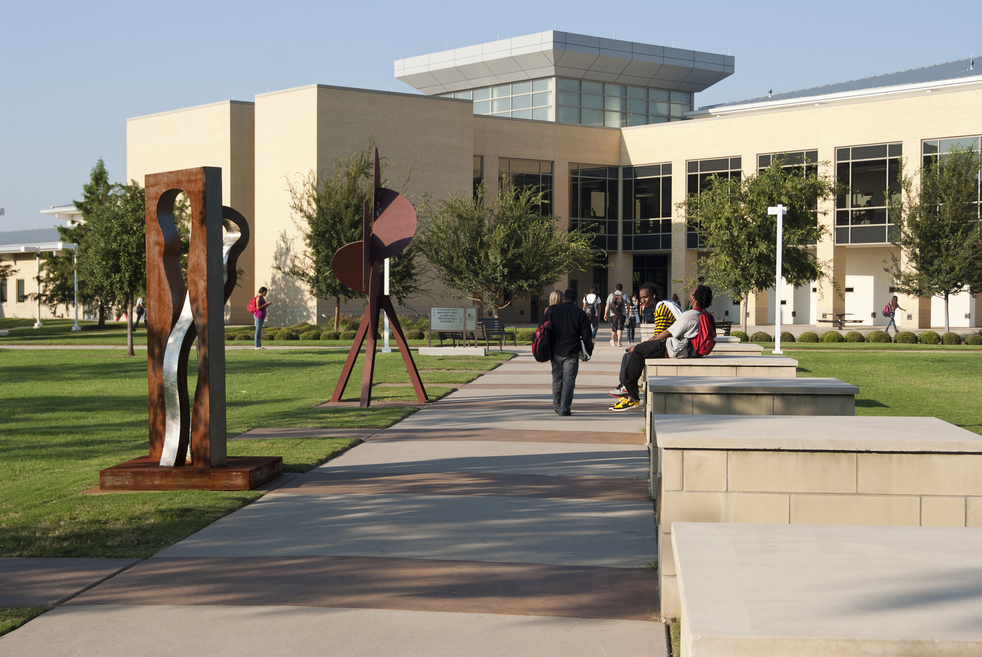 An outdoor photo of Collin College. Students walk down the sidewalk towards the main building. Art sculptures stand in the grass.