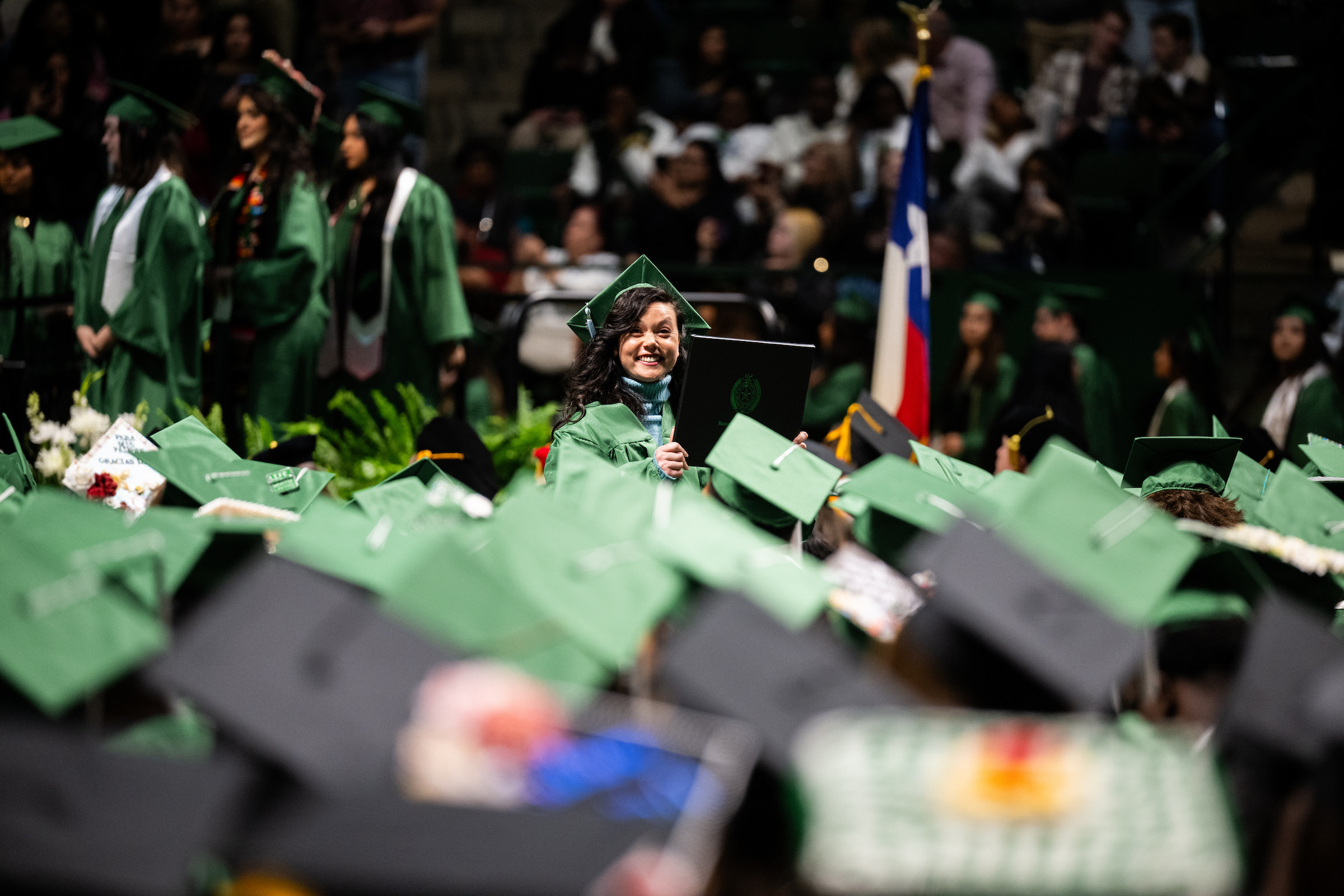 A young woman in her green graduation cap and gown walks past multiple students wearing green and black graduation caps. She is smiling at the camera and holding up her degree.