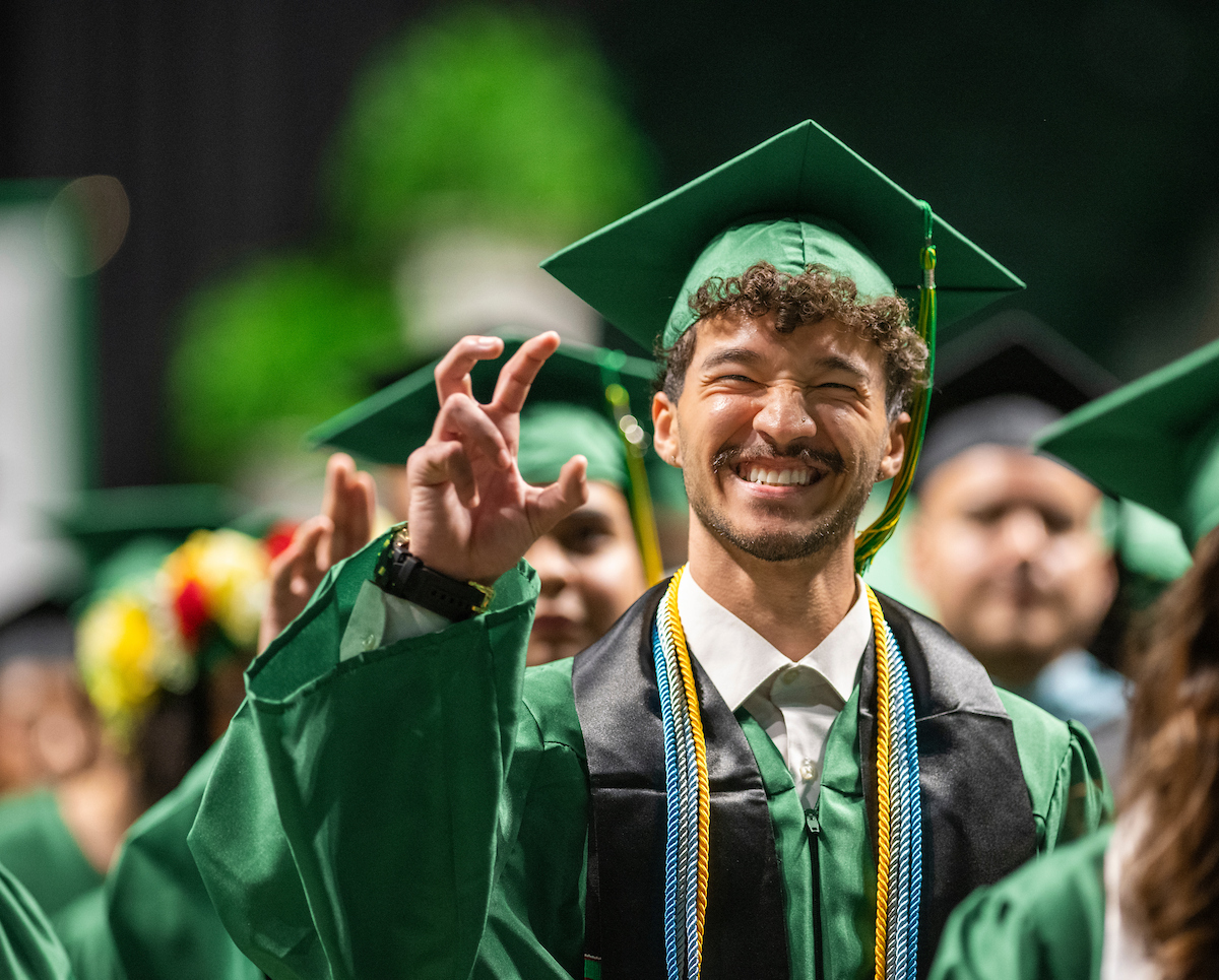 A male student in green graduation regalia looks at the camera with a big grin on his face. He makes the claw hand sign.