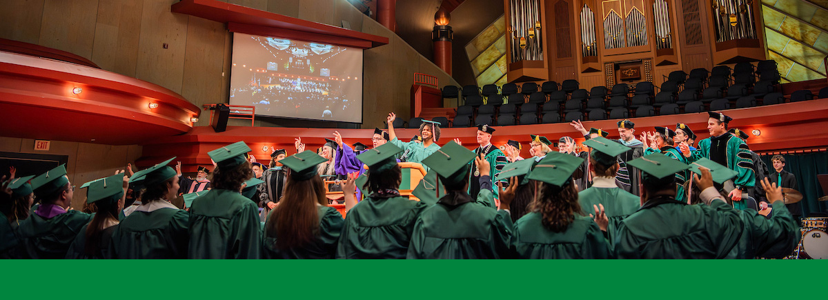 A group of students in green graduation regalia looks up at another student at a podium making the claw hand sign. A group of professors and President Keller stand behind them. Above is an organ on a balcony.
