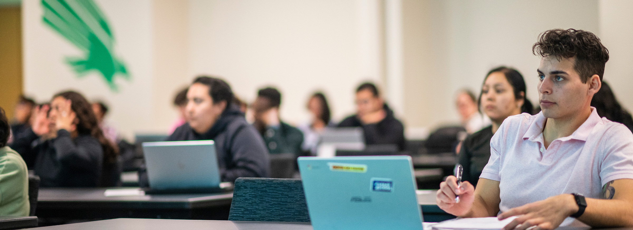 A photo of a male student sitting at a table with a laptop looking at something out of frame. He appears to be in class taking notes