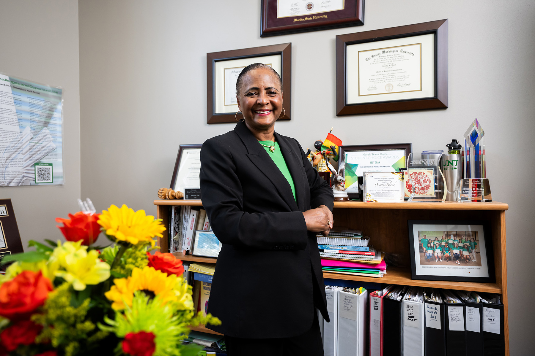 A woman stands in front of a bookcase wearing a black suit and green shirt