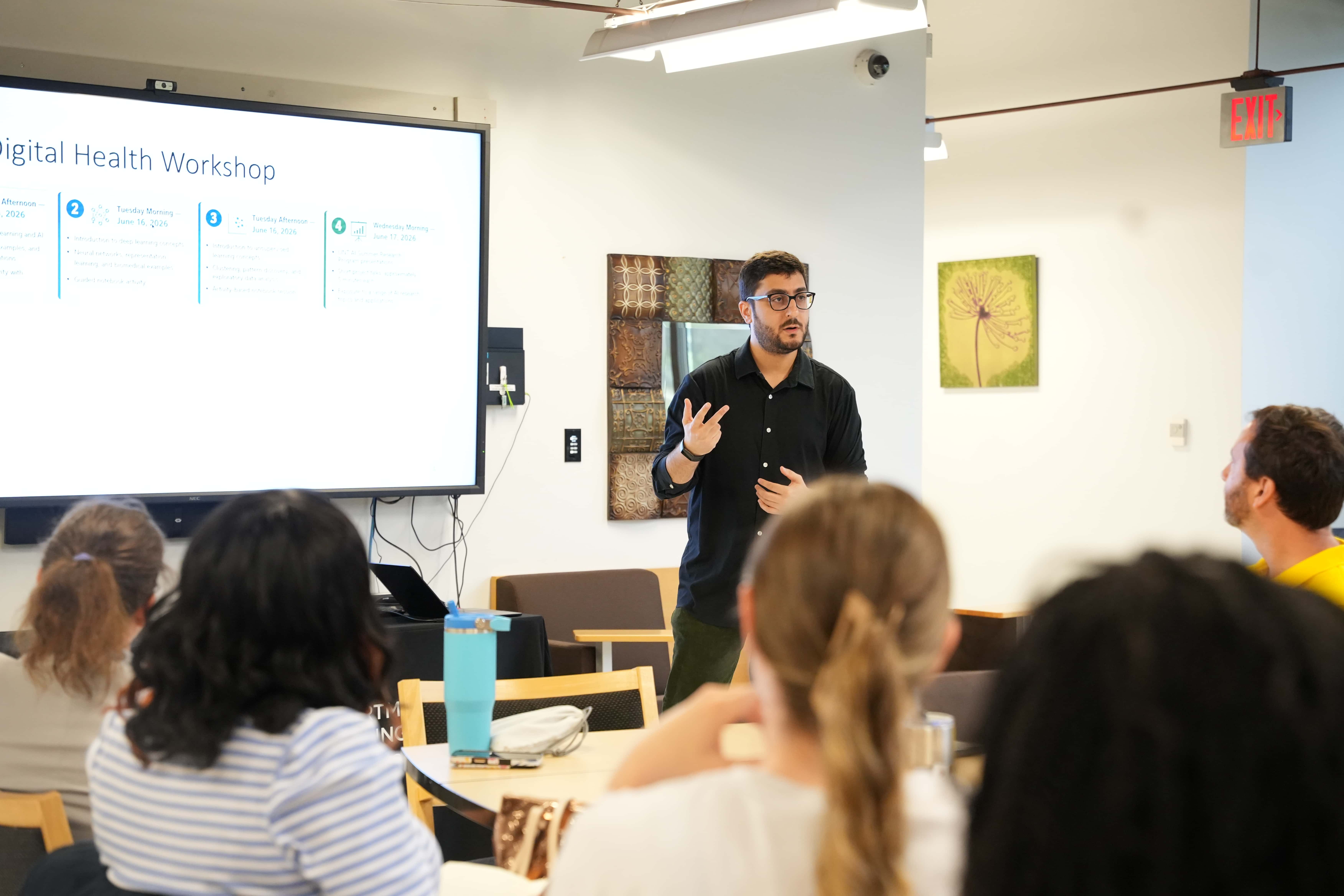 A man in a black button down stands before a group sitting at a table. Behind him, a projector reads "Digital Health Workshop"