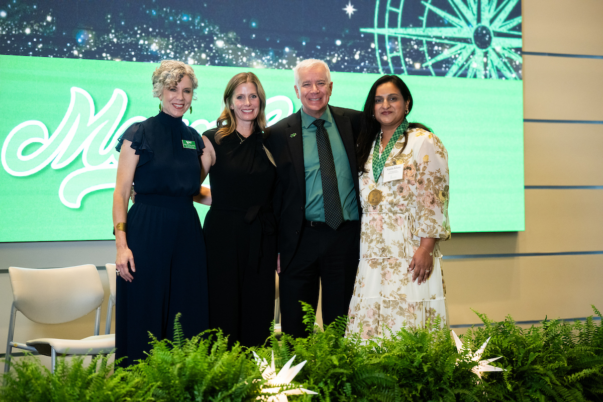 Two women, a man, and another woman side by side posing for a photo. The first two wear all back, the man wears a green shirt with black pants and blazer, the final woman, Nazia Khan, wears a white dress and a medal.