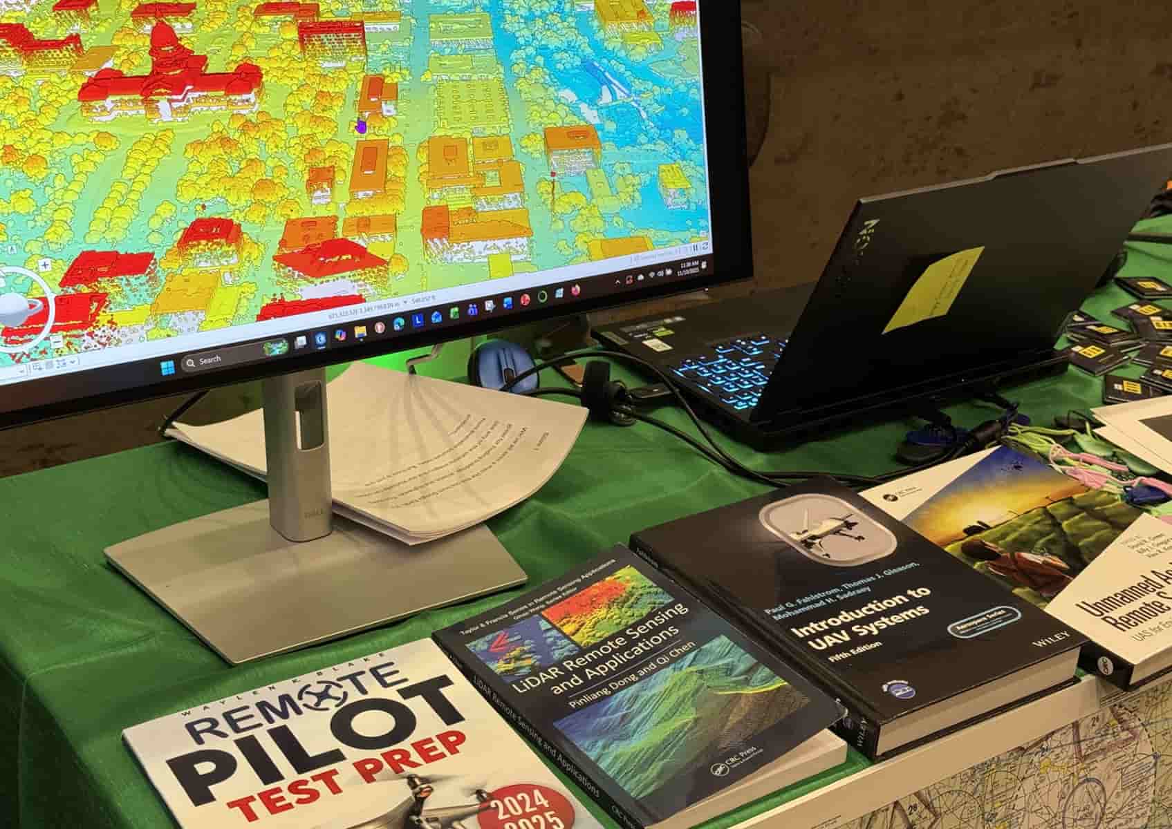A computer monitor displays colorful LIDAR imagery. Below the screen are a row of books on remote piloting and drones.