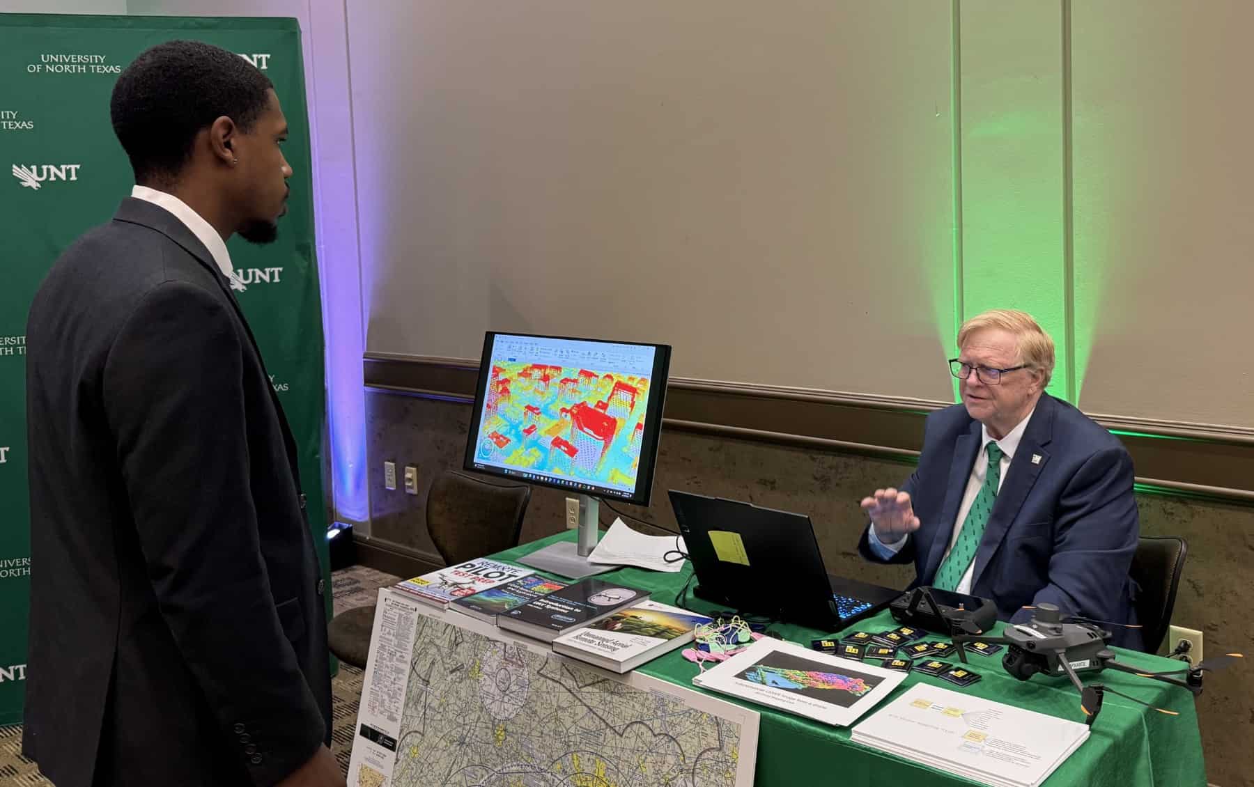 A man stands in front of a table of books and a computer monitor, behind which another man is seated. 