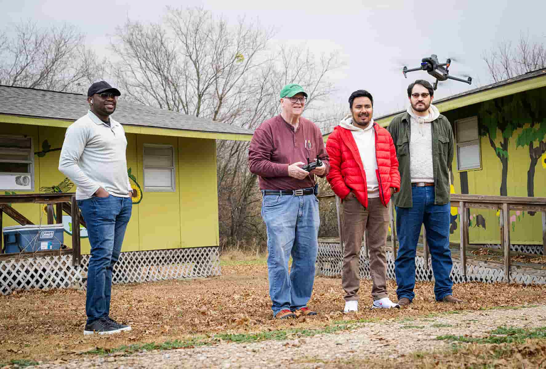 Four people stand outside in front of yellow buildings as one person pilots a drone using a handheld controller.