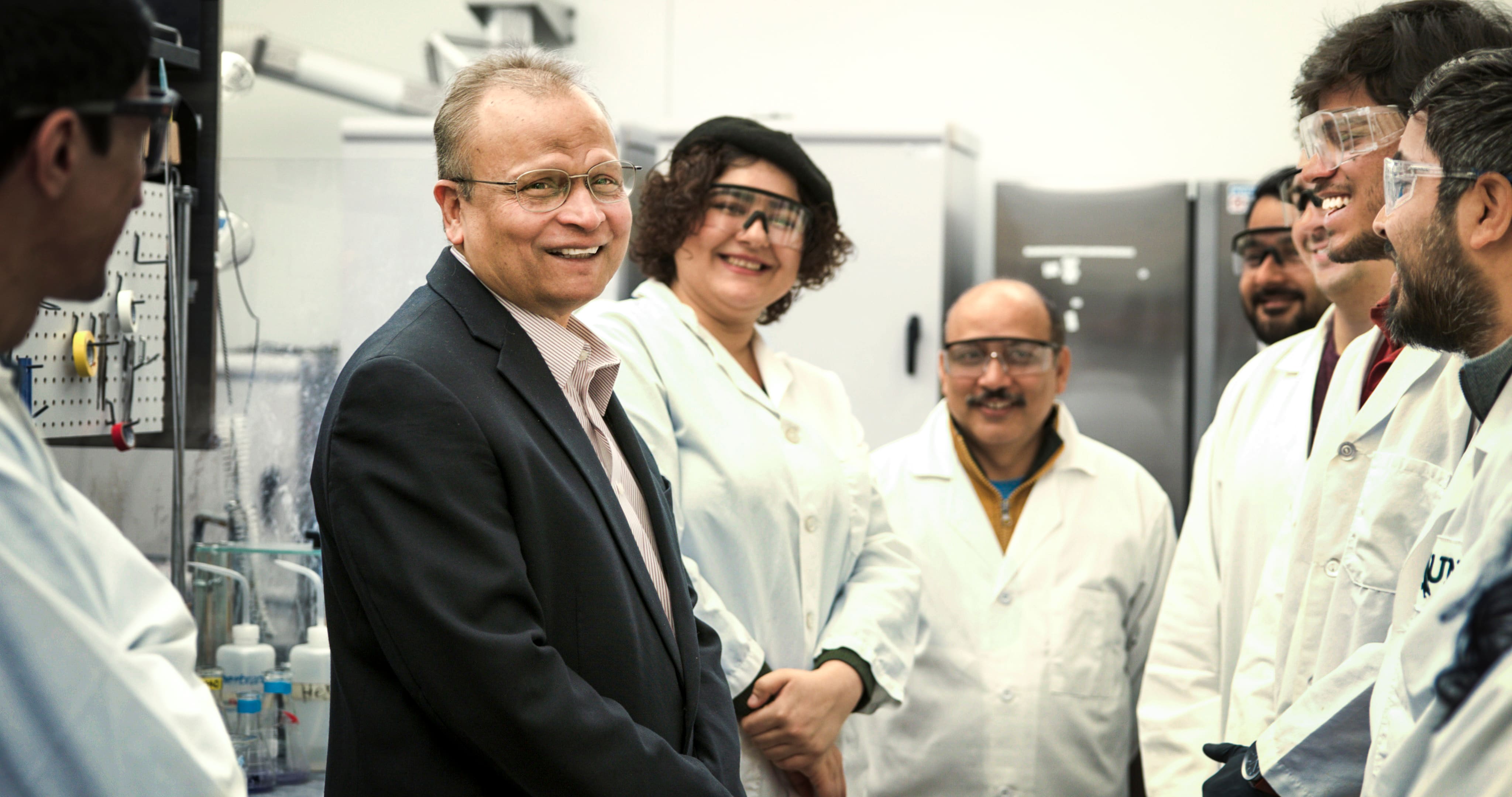 Francis D'Souza wearing a suit stands in a lab surrounded by his students in lab coats. They're all smiling