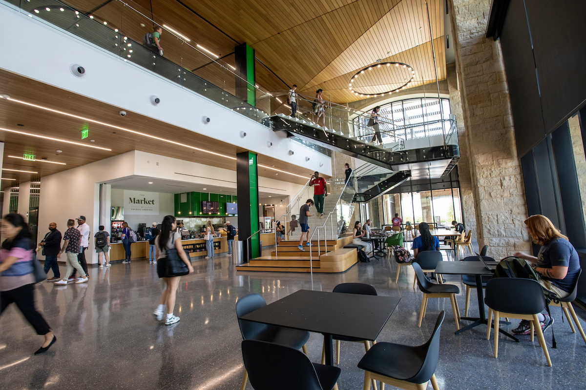 A photo of the lobby of UNT at Frisco. Students sit at the tables, walk by the cafe, and walk up the stairs. A green pillar is in the middle with a market open to the left.