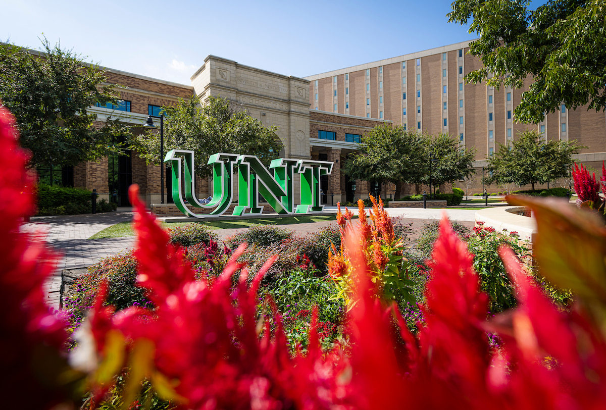 A photo of the green UNT letters statue with red flowers in the foreground.