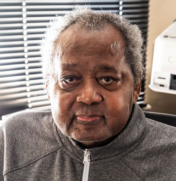 An older man wearing a gray zip up jacket looks at the camera with a neutral expression.