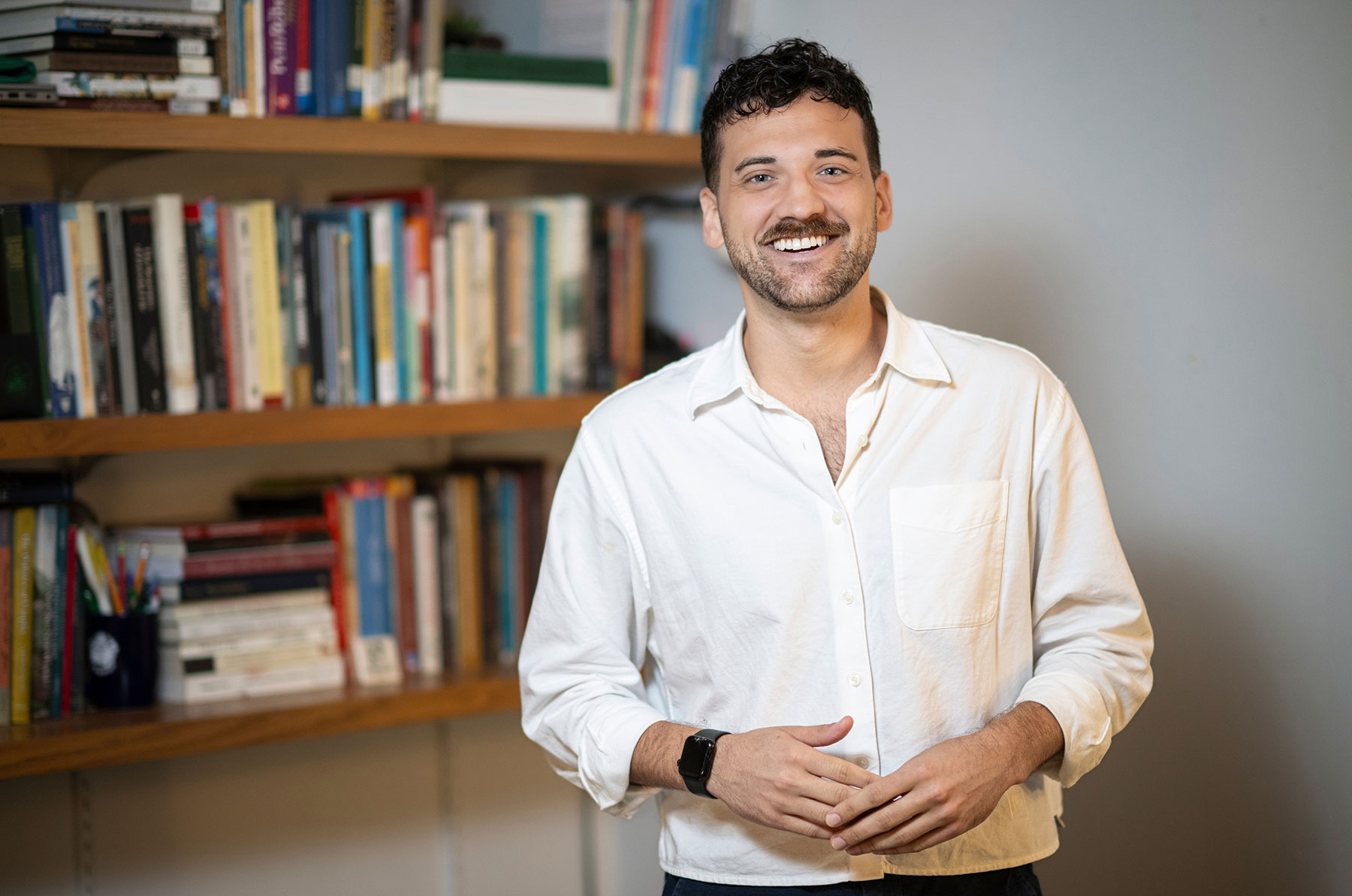 Jakob Burnham stands in front of a bookshelf. He wears a white button down shirt.