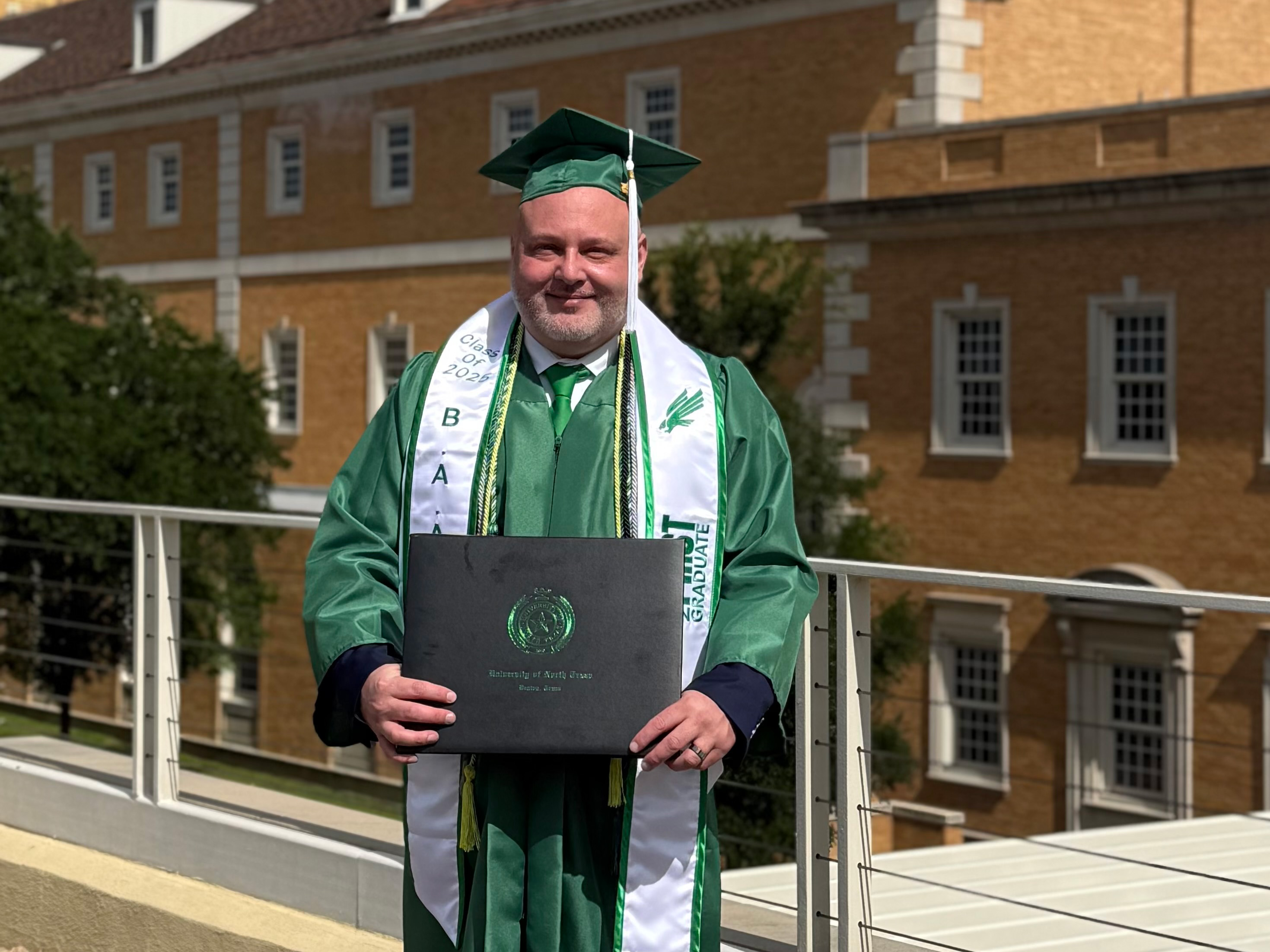 John Iglehart-Fernandez, smiling, stands in green cap and gown holding his degree in front of a UNT building