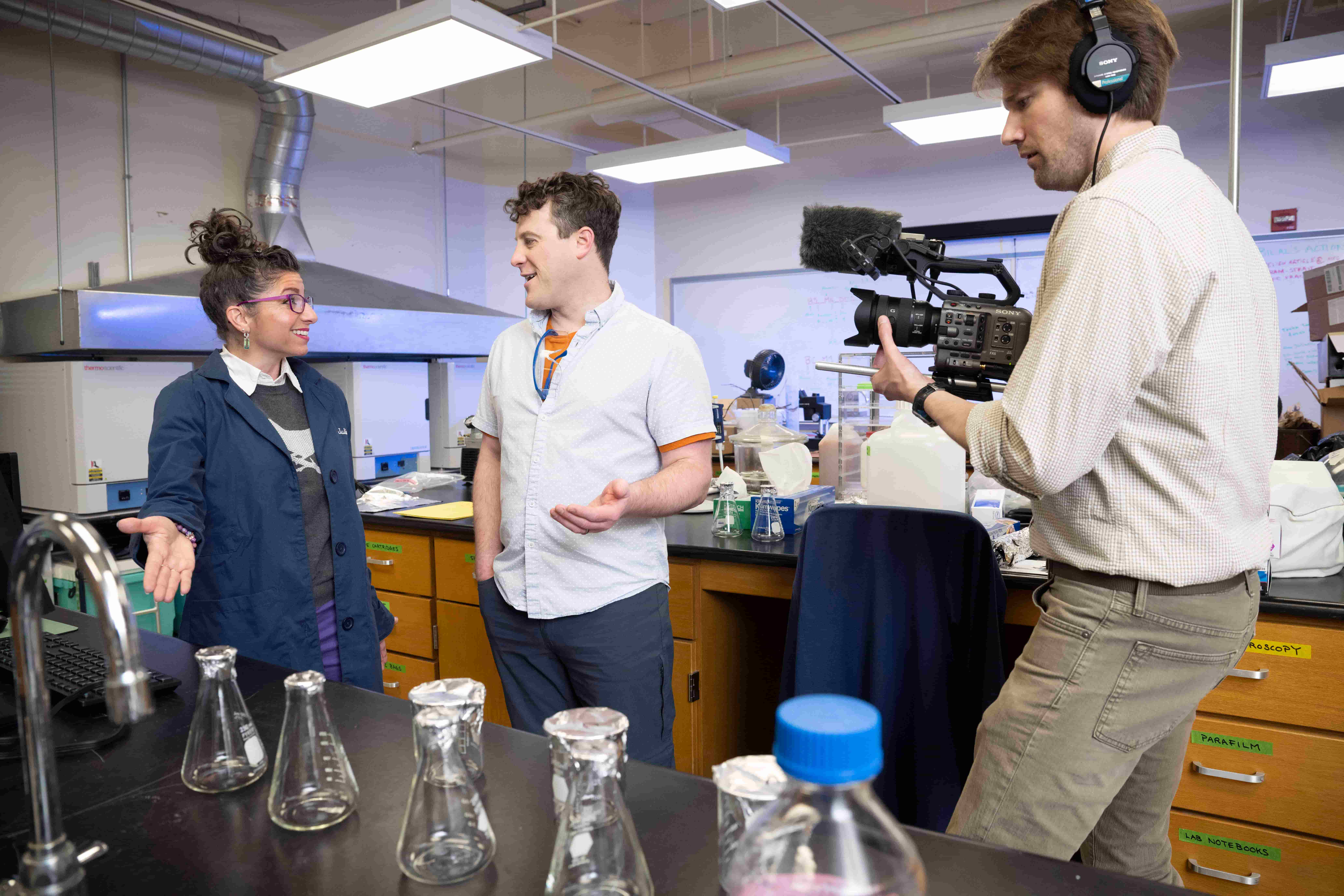 Juliana D'Andrili and Jim Junker stand talking to one another, Brandon Gaesser stands to the right filming them. All three are in a science lab.