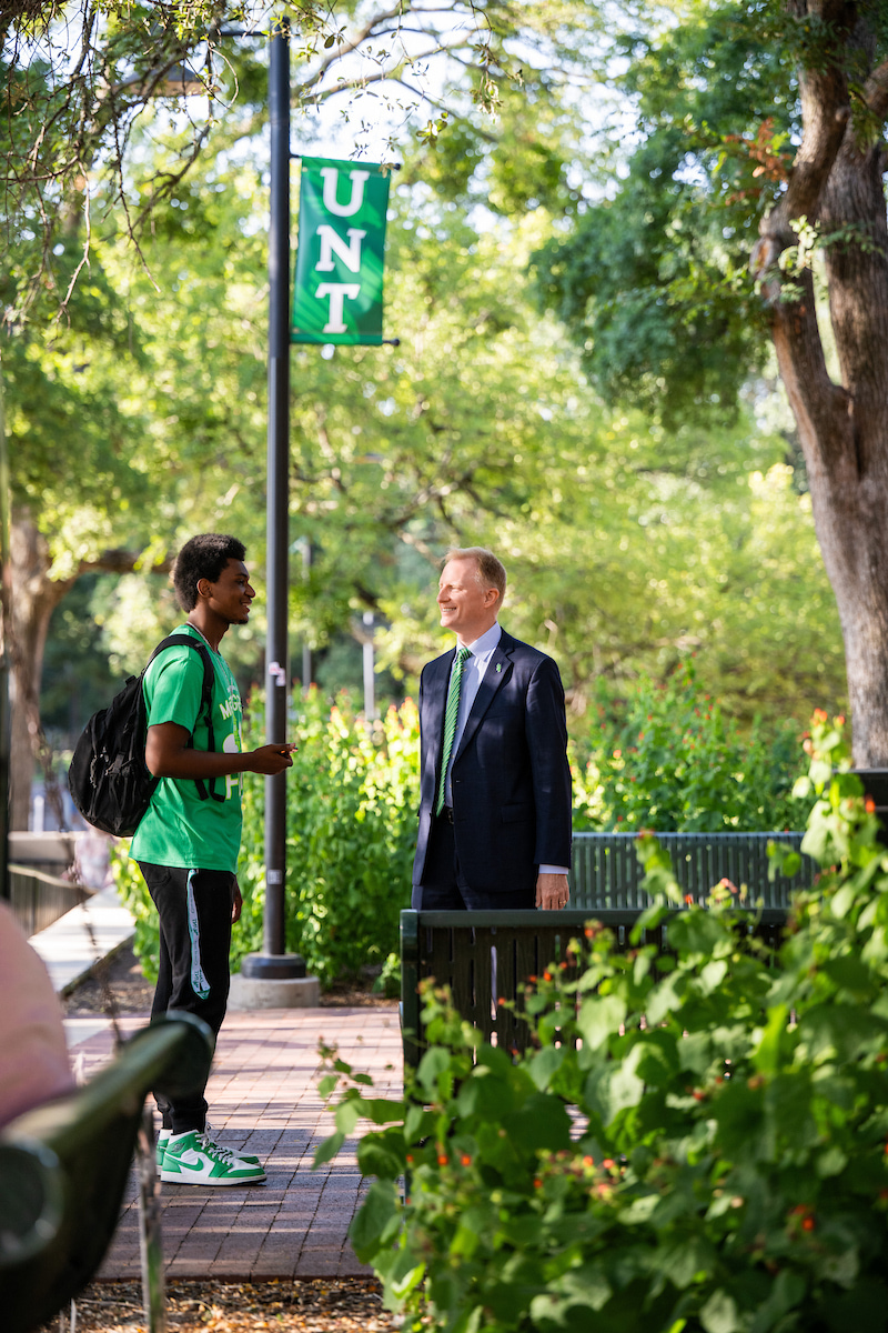 A young man wearing green stands left to President Keller standing right who is wearing a suit and green tie. They are talking beneath a flag pole with a UNT banner on it. They are surrounded by trees and shrubbery