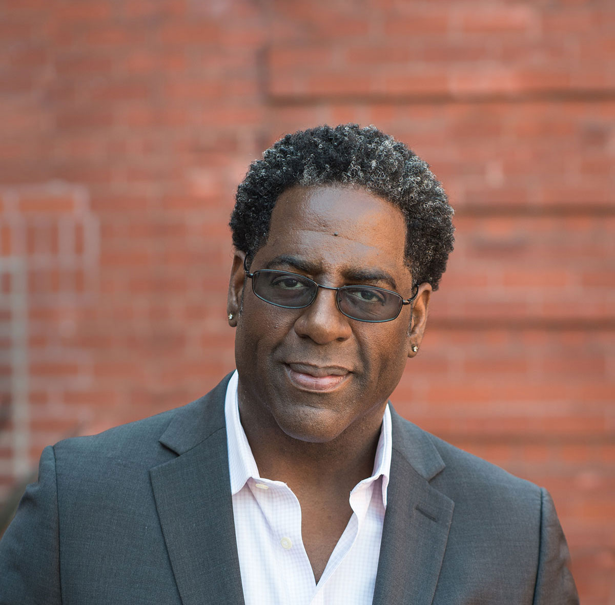 A man in a gray suit stands in front of a red brick wall