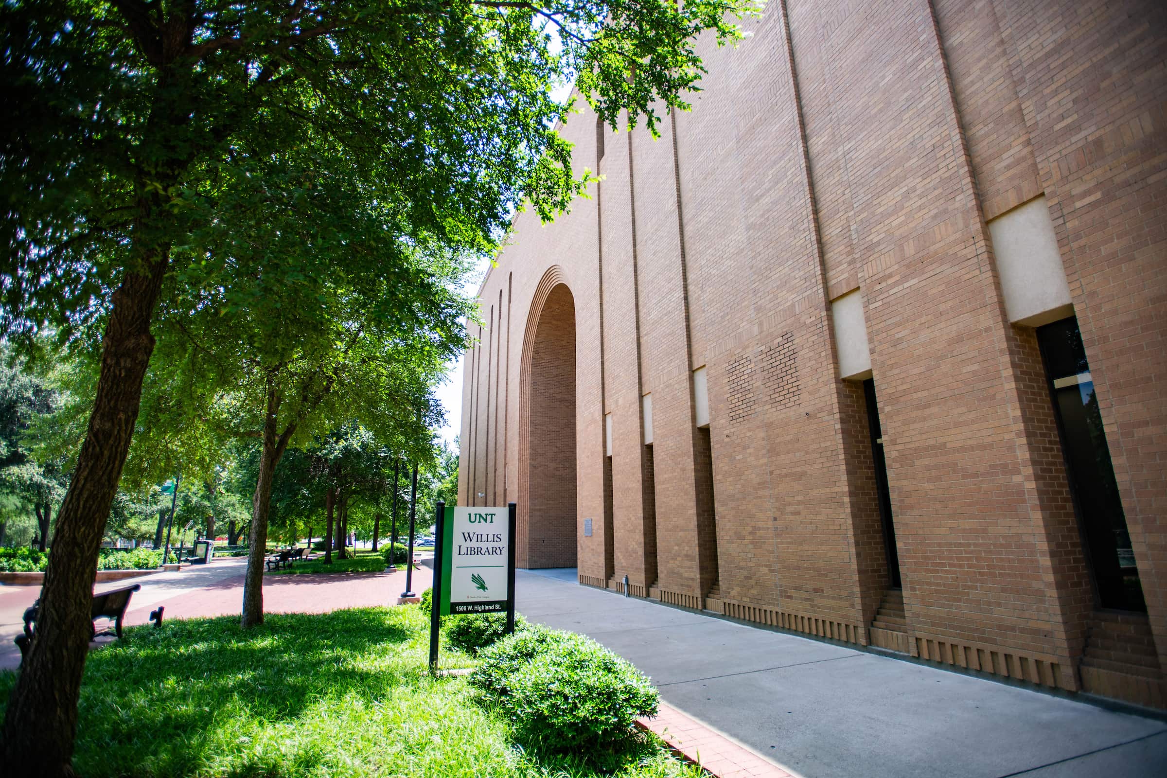 A photograph showing the front of Willis Library from a side angle. The building has brown brick walls with a sidewalk in front of it. Green trees and grass grow to the left