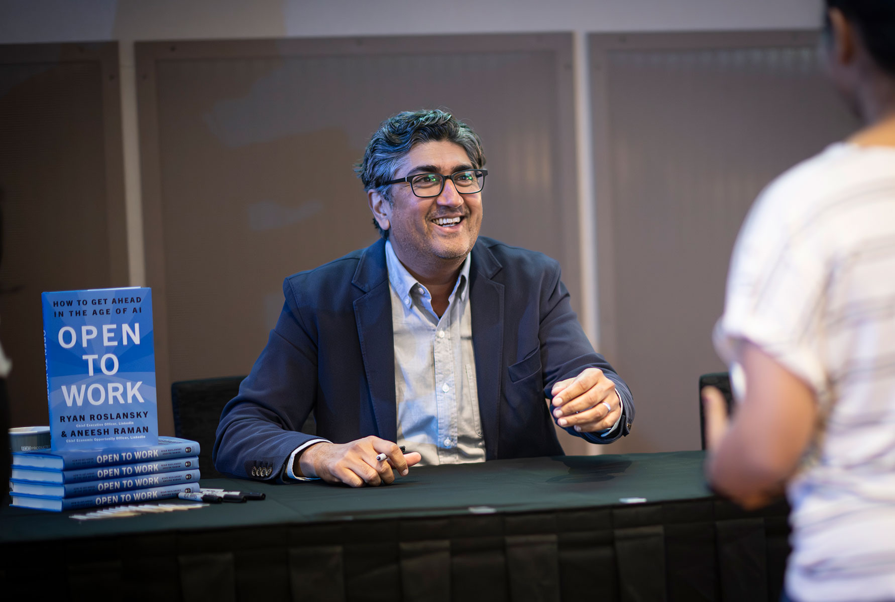 A man sits behind a table with a stack of books on it, speaking to a person who wants their book signed